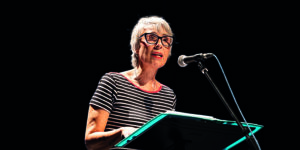Carmen García Nieto en un acto público, leyendo un discurso desde un atril con micrófono, vestida con una camiseta de rayas y gafas