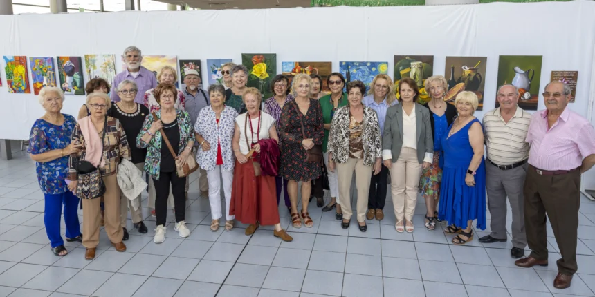 Grupo de mujeres junto al cartel de la exposición “La Magia del Color” en el Centro Municipal de Ocio de Torrevieja, con pinturas al fondo.